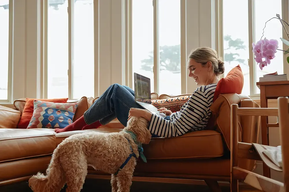 Femme assise sur un canap&eacute; avec un chien &agrave; ses pieds, travaillant sur un ordinateur portable pr&egrave;s d'une fen&ecirc;tre lumineuse.